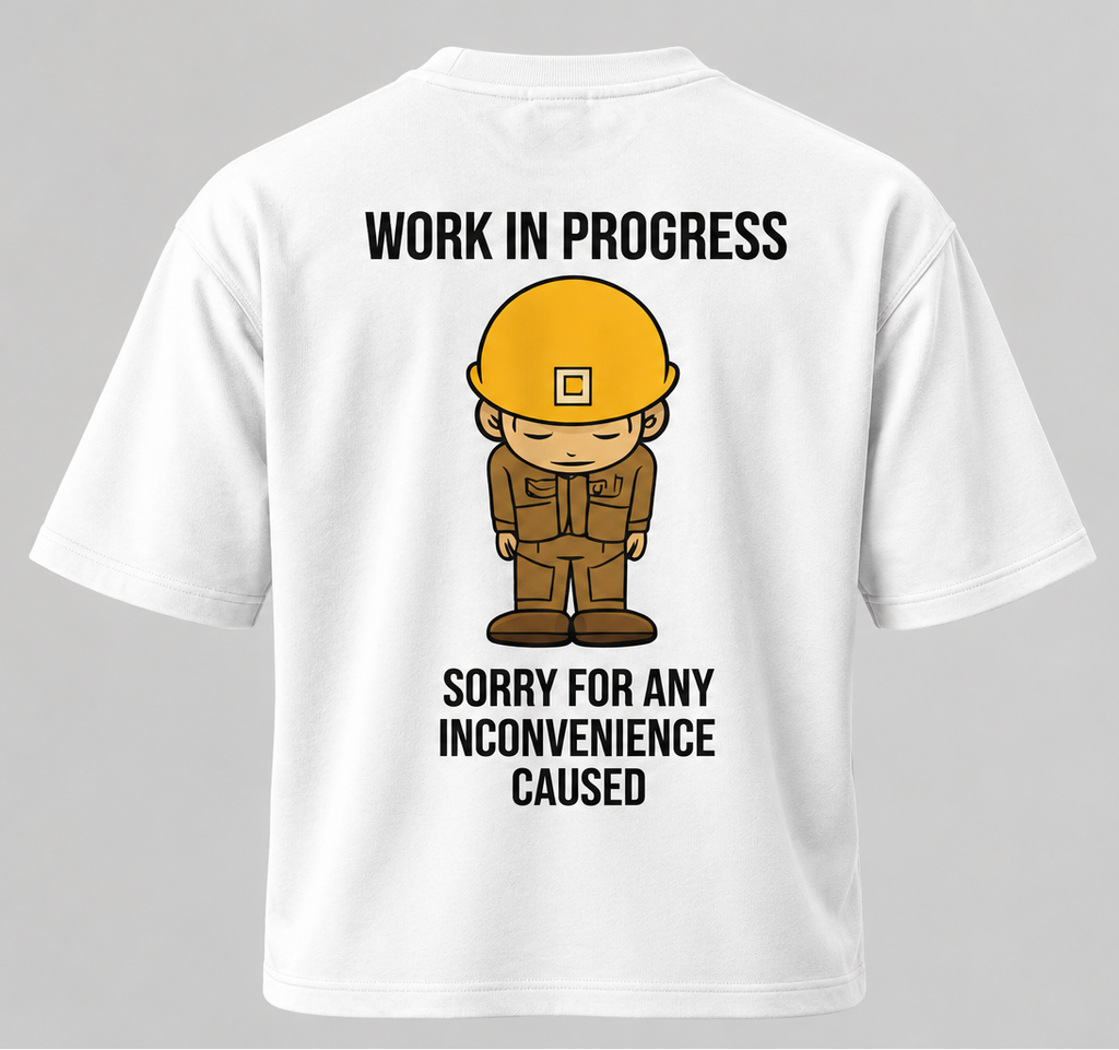 White short-sleeve t-shirt shown from the back, featuring a cartoon construction worker graphic with a yellow hard hat and the text 'WORK IN PROGRESS' above and 'SORRY FOR ANY INCONVENIENCE CAUSED' below.