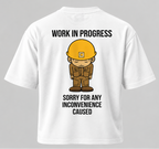 White short-sleeve t-shirt shown from the back, featuring a cartoon construction worker graphic with a yellow hard hat and the text 'WORK IN PROGRESS' above and 'SORRY FOR ANY INCONVENIENCE CAUSED' below.